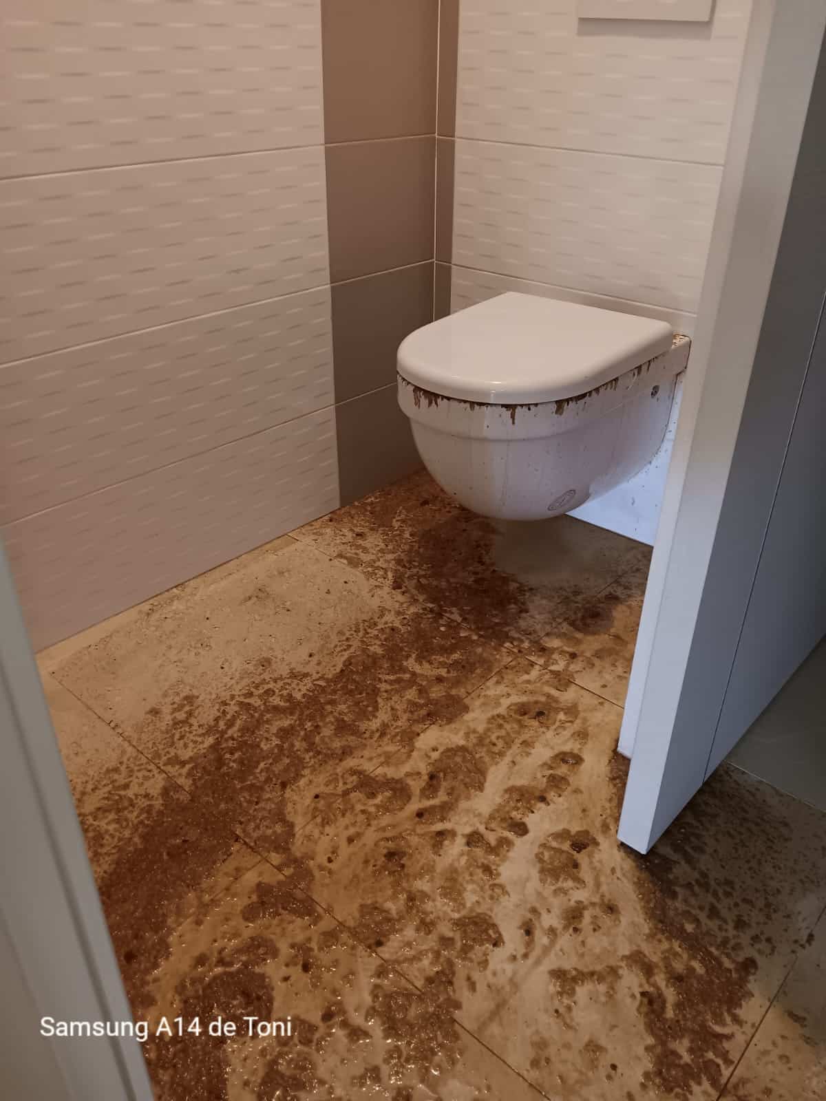 Toilettes modernes recouvertes et sol de salle de bain inondé de matières brunes. Texte visible: Samsung A14 de Toni.
