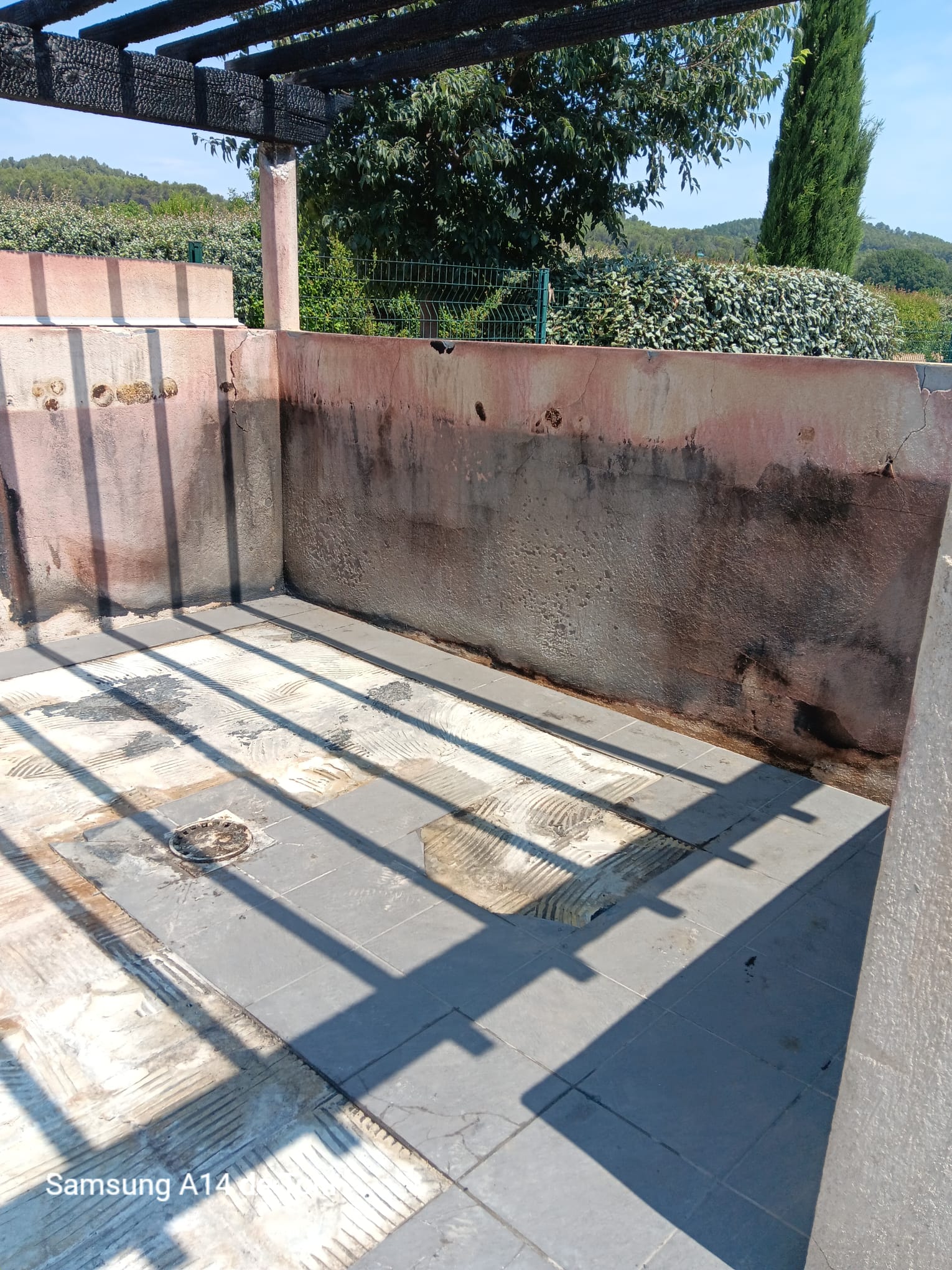 Bassin vide avec carrelage arraché, murs abîmés et pergola noircie par le feu ; texte "Samsung A14" visible.