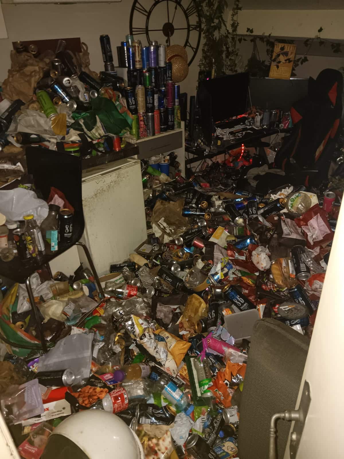 Pièce jonchée d'une quantité massive de canettes de boissons énergisantes 86, bouteilles et emballages alimentaires, désordre extrême.