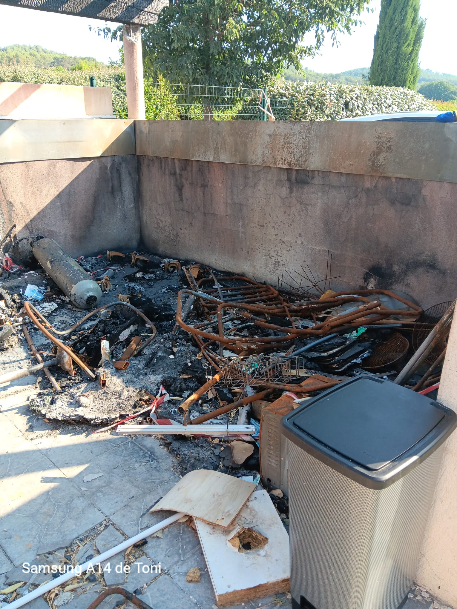 Décombres calcinés et rouillés d'un incendie dans un espace extérieur dallé, avec une grande bouteille de gaz. Texte visible : Samsung A14 de Toni.