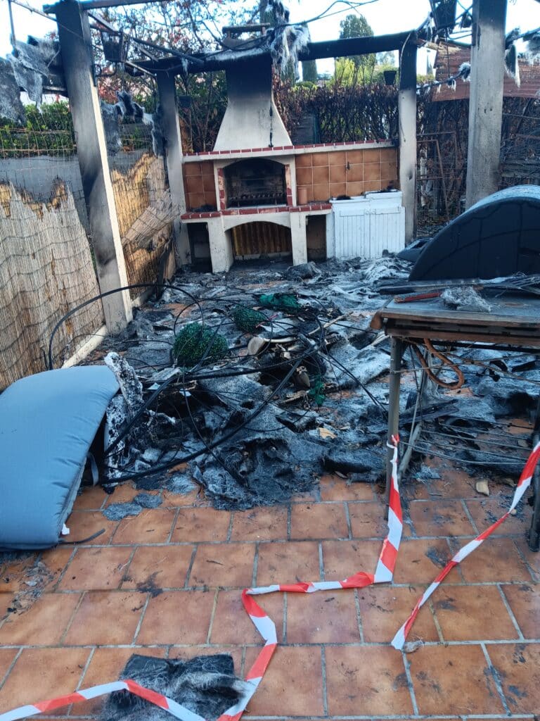 Terrasse extérieure sinistrée par un incendie : débris brûlés, structure noircie, ruban de signalisation rouge et blanc, barbecue en arrière-plan.